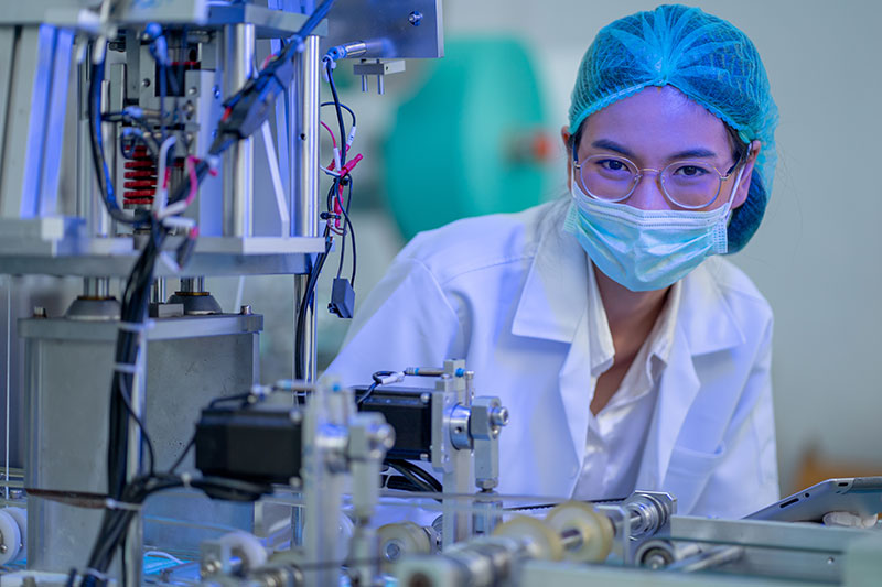 Factory worker woman with gown uniform stand near part of mask producing machine