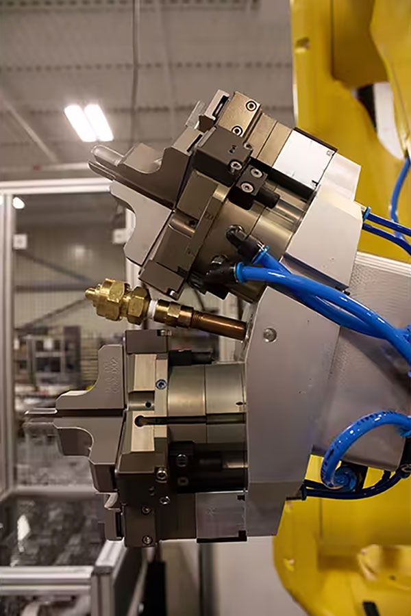 An industrial robotic arm with a dual pneumatic gripper tool and blue air hoses installed in manufacturing facility with safety enclosures in the background.