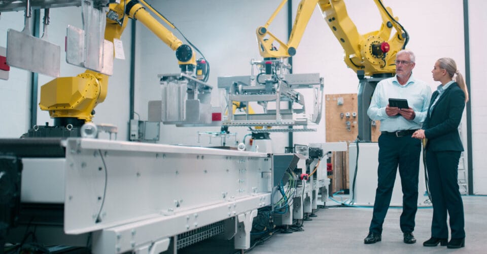 A man and woman observe two robotic systems working on a manufacturing production line.