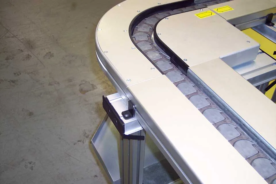 The rounded corner of a Tech-Track conveyor system with a white metal housing and silver conveyor belt.